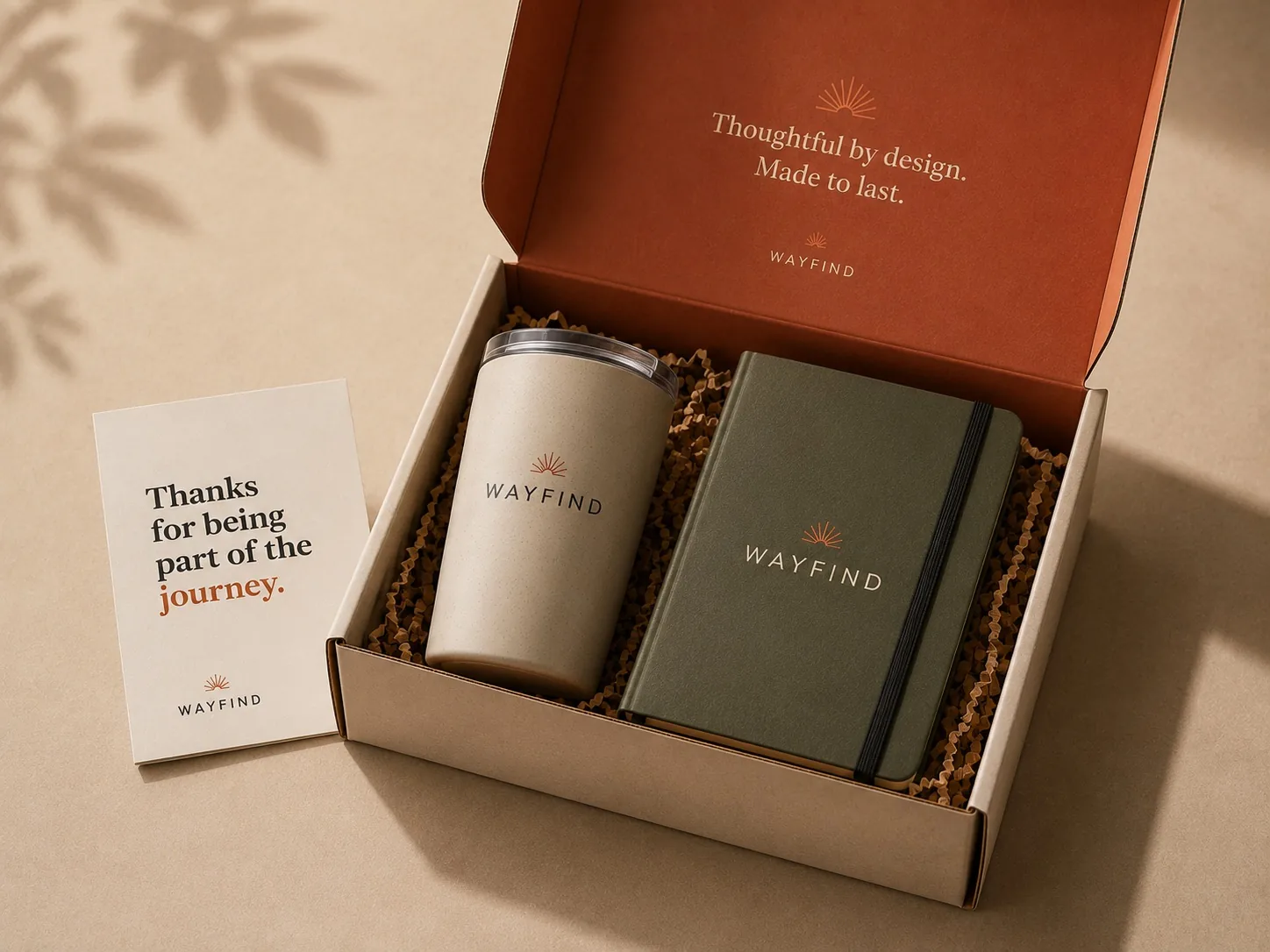An open gift box holding a branded tumbler and notebook on kraft crinkle fill, beside a thank-you card.